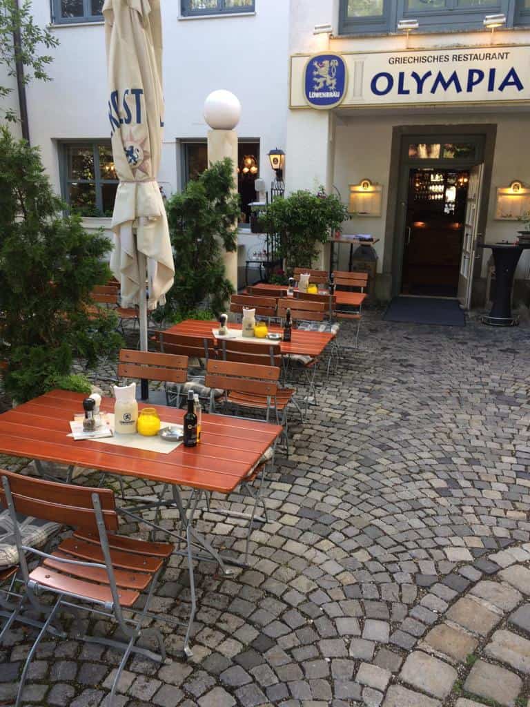 Griechischer Biergarten des Restaurant Olympia mit Gästen an Holztischen unter Olivenbäumen